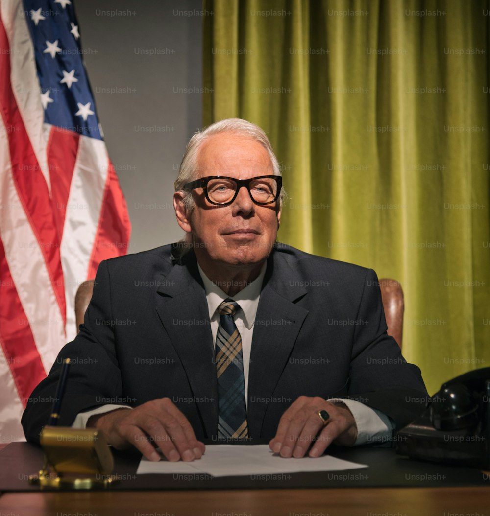 Big Boss Präsident mit Brille, sitzt hinter dem Schreibtisch mit amerikanischer Flagge im Hintergrund.