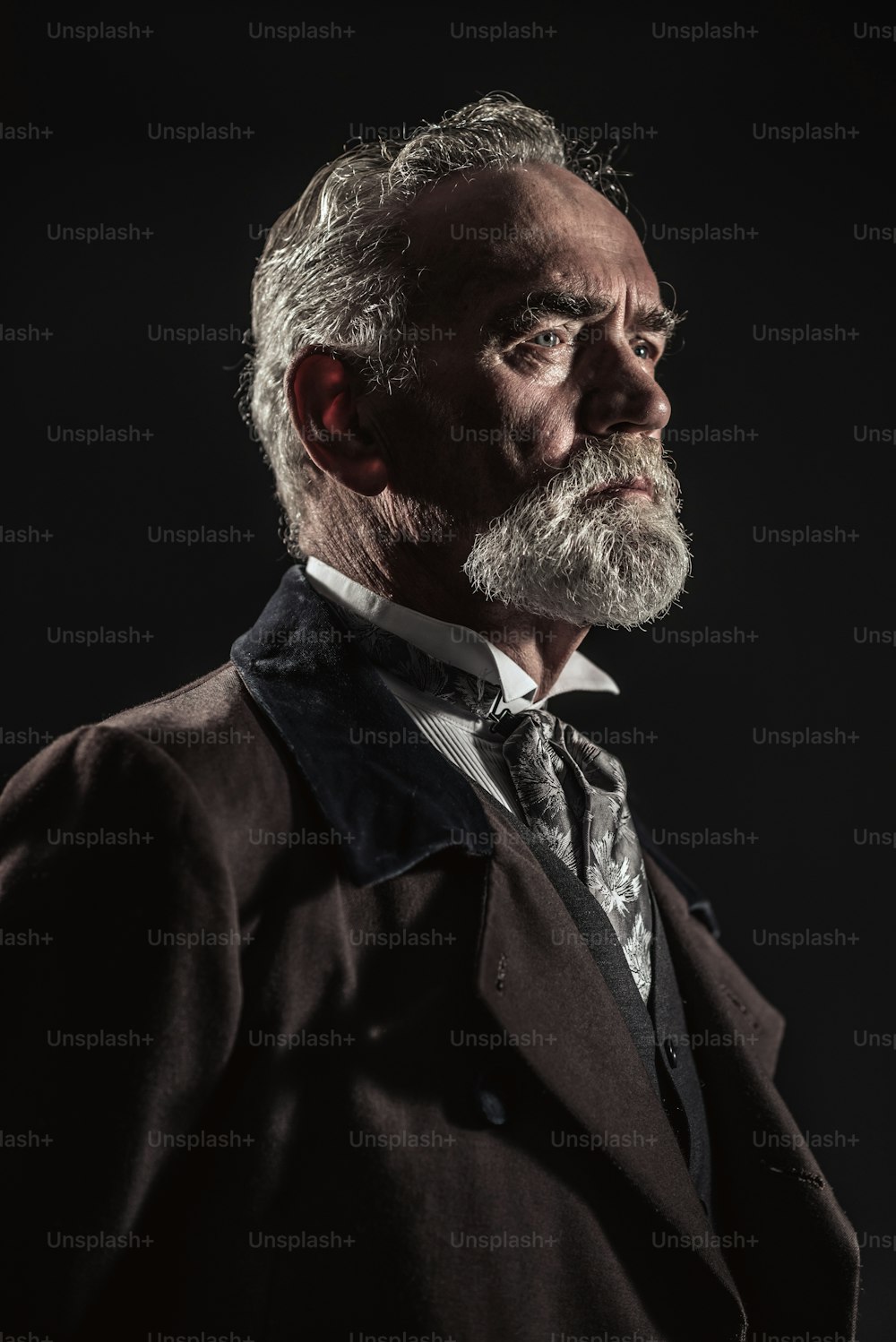 Hombre mayor de características vintage con cabello y barba grises. Estudio filmado sobre fondo oscuro.