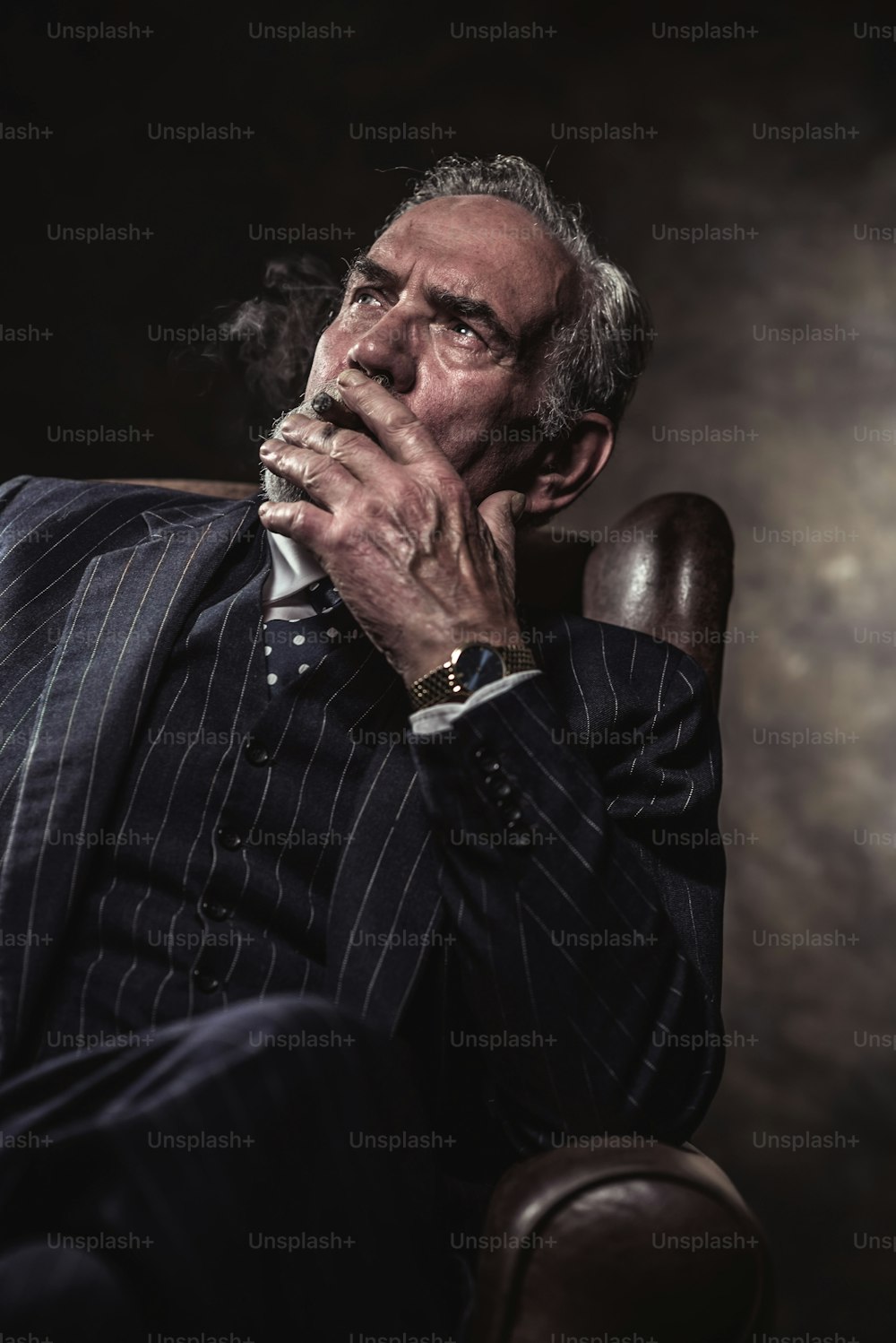 Sulla sedia seduto caratteristico uomo d'affari senior. Fumare sigaro. Capelli e barba grigi che indossano abito e cravatta a righe blu. Contro muro marrone.
