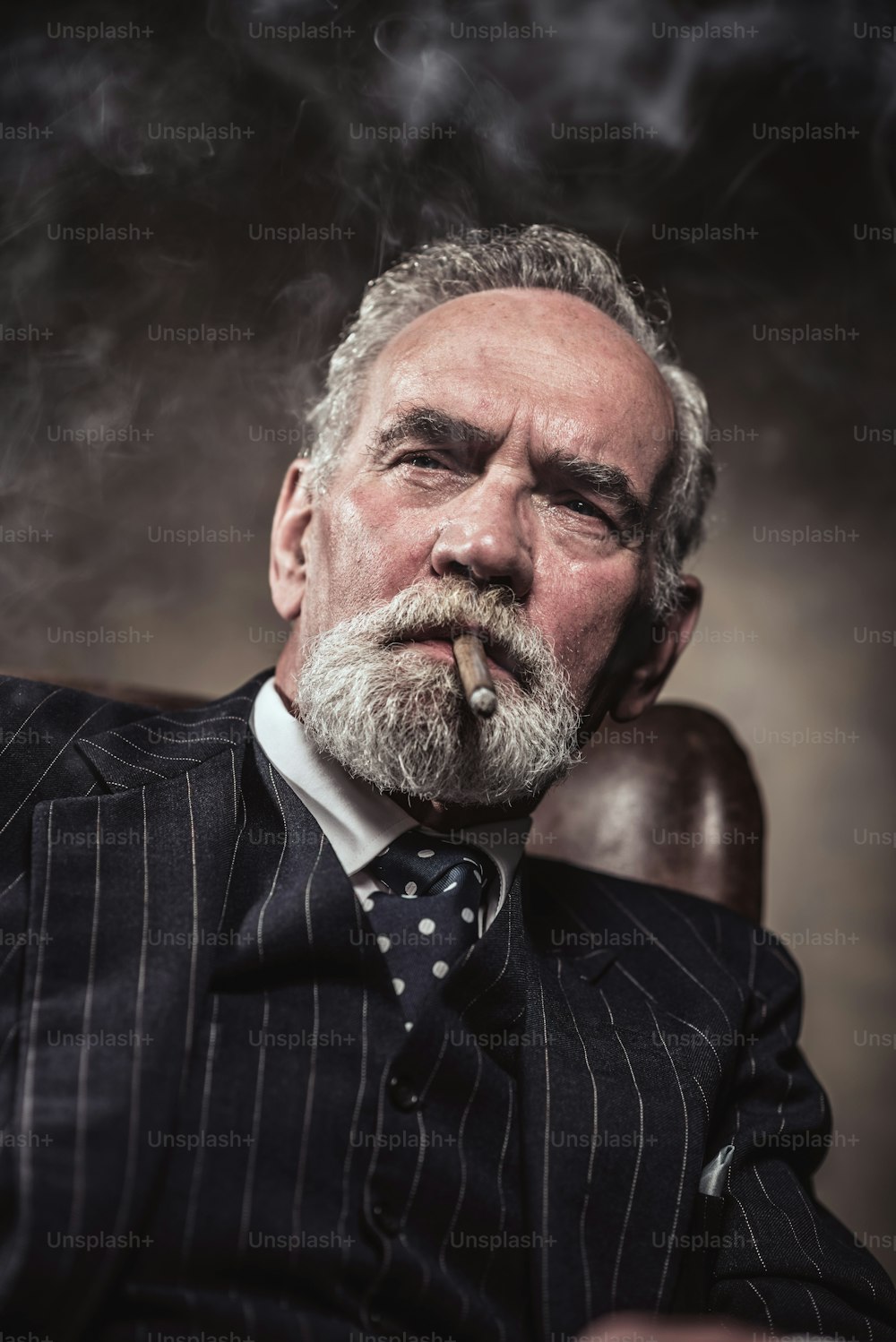 Sulla sedia seduto caratteristico uomo d'affari senior. Fumare sigaro. Capelli e barba grigi che indossano abito e cravatta a righe blu. Contro muro marrone.