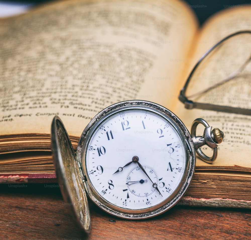 木のオフィスデスクの背景にビンテージ懐中時計と古い本を開く、接写。時間、科学の概念