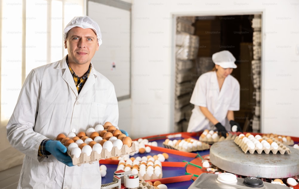 Giovane agricoltore maschio europeo in uniforme che tiene la scatola di cartone piena di uova di gallina fresche dopo la cernita e l'etichettatura nell'allevamento di pollame