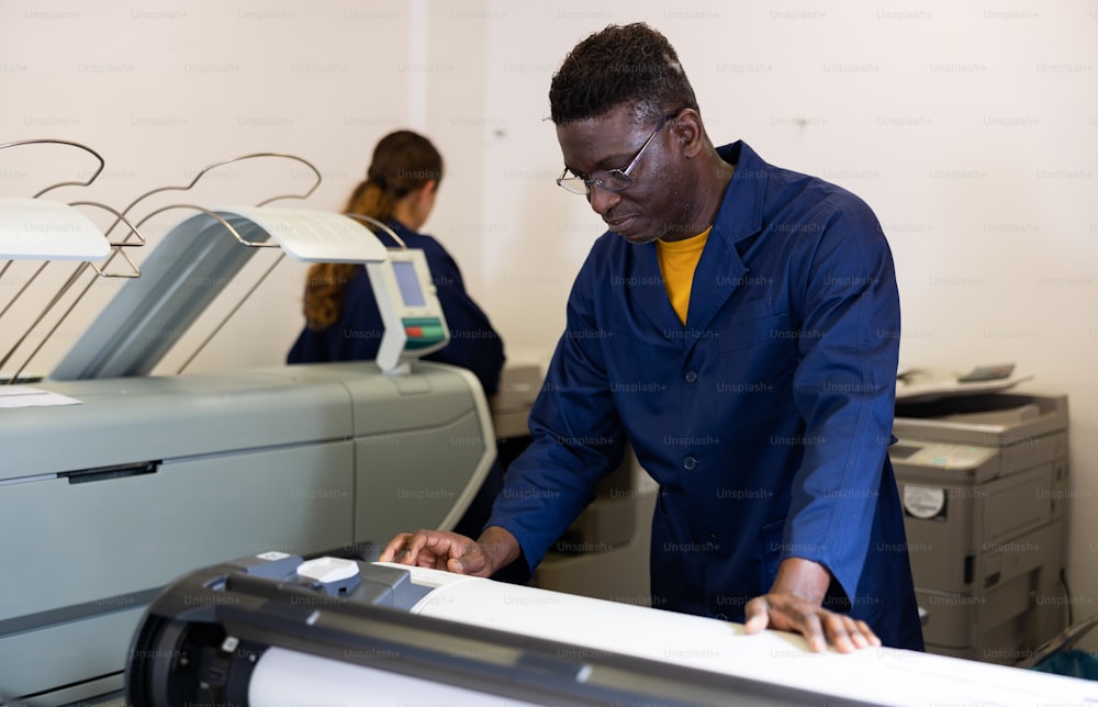 Homme afro-américain d’âge moyen positif en uniforme utilisant un traceur pendant le travail à l’imprimerie