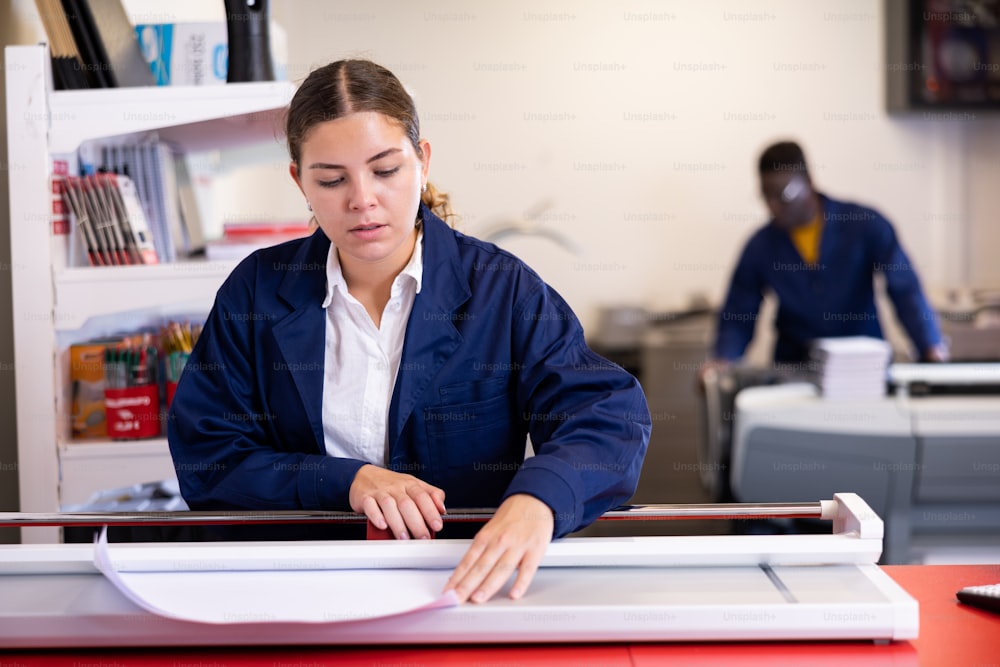 Mujer joven seria con un uniforme azul usando un cortador de papel en la mesa con planificadores y calculadora en la imprenta