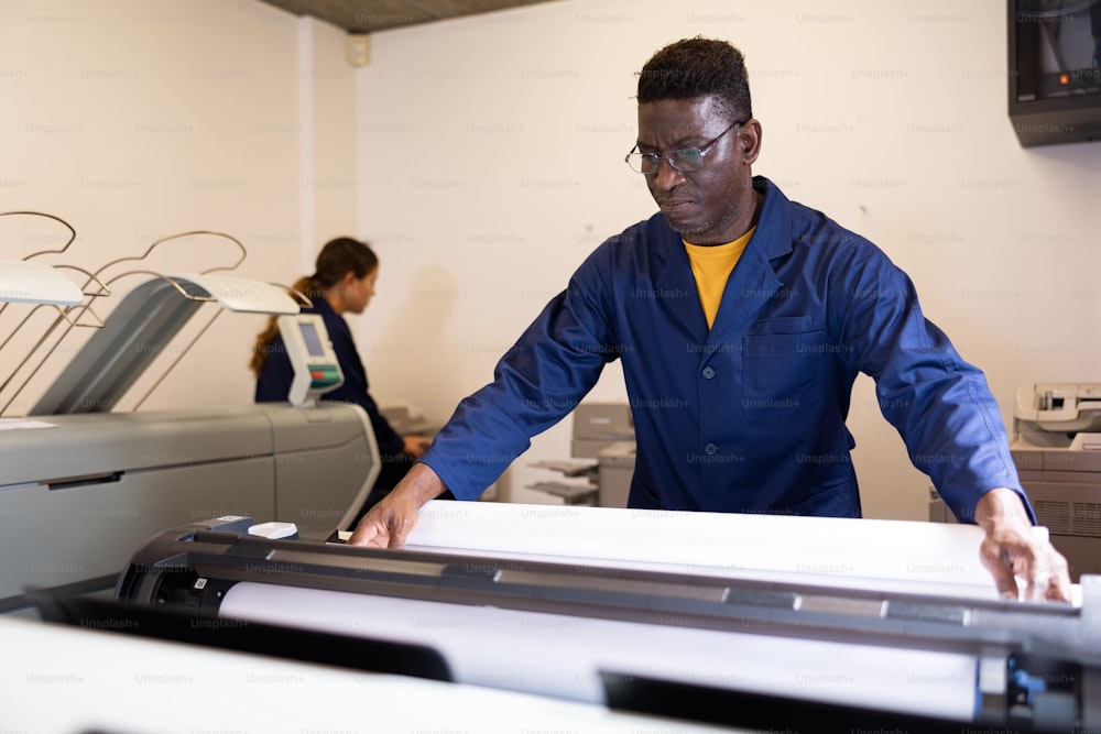 Hombre afroamericano serio de mediana edad en uniforme cargando papel de gran formato en un plotter en la imprenta