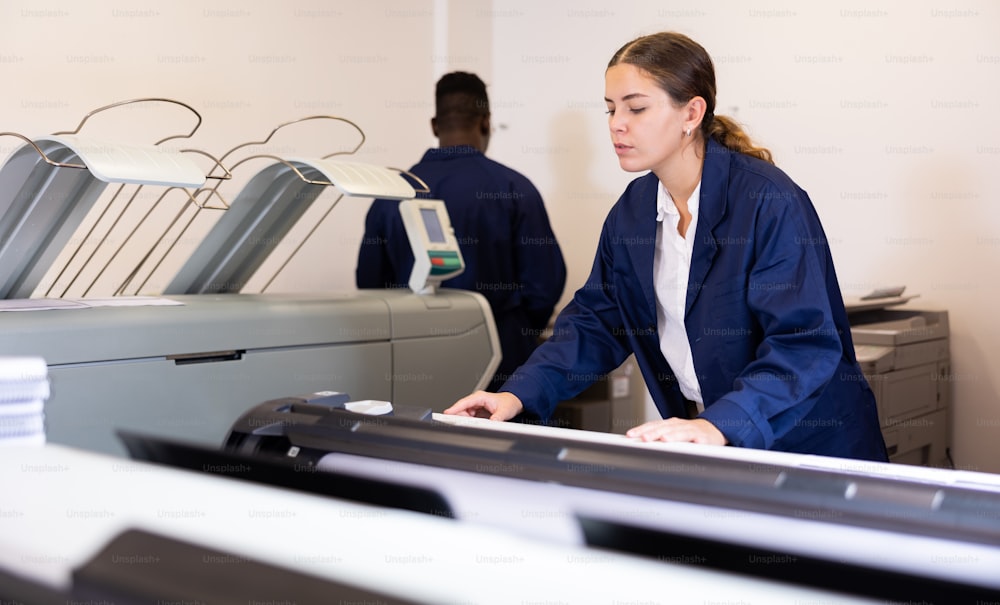 Focused young woman in uniform setting up the plotter before starting work in the printing house