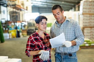 Caucasian man and latin woman co-workers talking about stock control in warehouse, verifying check list.