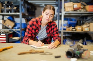 Portrait of young woman storehouse worker standing at workbench and writing, making notes.