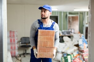 Portrait of a young man builder in work overalls carrying bricks during finishing work indoors