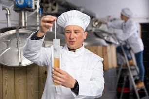 Interested brewery owner checking alcoholic content in craft beer, using test tube and hydrometer while standing near fermenters in workshop