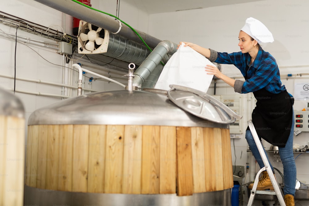 Professional young female brewer working in small brewery, pouring malted grain from bag into fermenter to produce beer