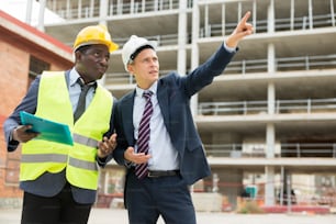 Afroamerikanischer Architekt und europäischer Ingenieur in Warnvest diskutieren über das Projekt eines neuen Gebäudes. Ein Mann zeigt mit dem Finger, ein anderer hält die Projektdokumentation in der Hand.