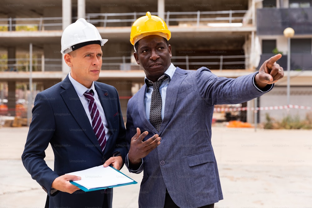 African-american and Caucasian architects standing on building site and discussing new construction. One of them pointing with finger.