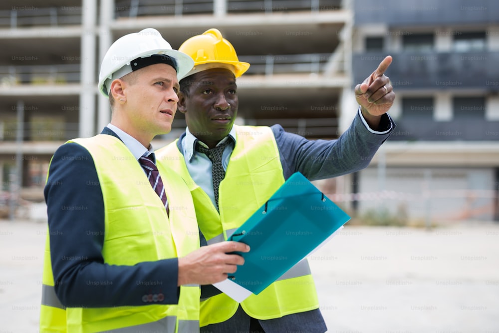 African-american and European men standing on construction site and talking. One of them holding folder with documentation, another one showing something with finger gesture.