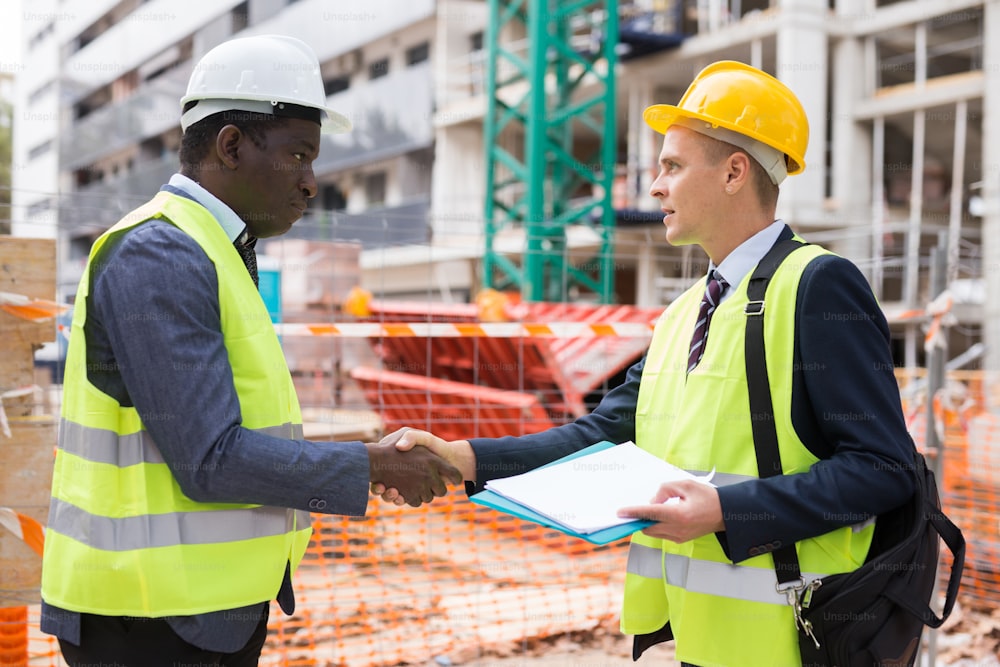 Two successful civil engineers at a construction site exchange a friendly handshake, holding documents in their hands