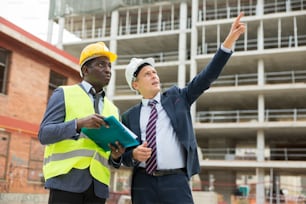 African-american architect and European engineer in warnvest discussing project of new building. One man showing with finger, another one holding project documentation.