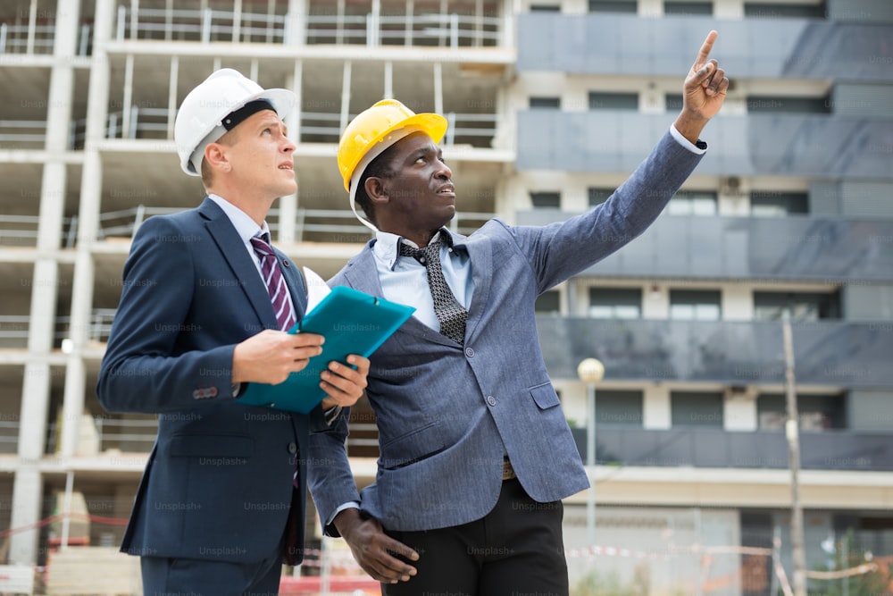 African-american and Caucasian architects standing on building site and discussing new construction. One of them pointing with finger.