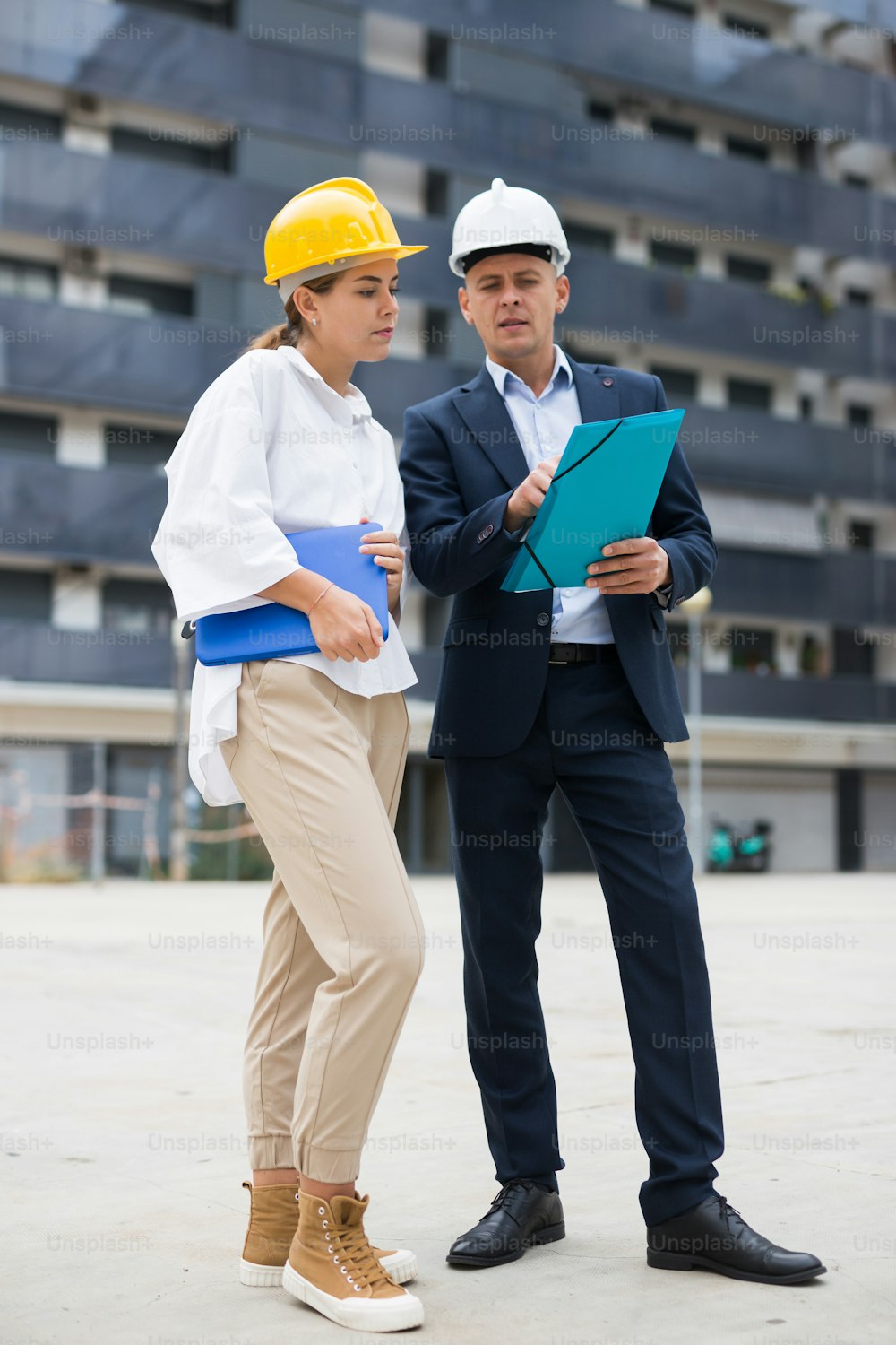 Two young successful architects, controlling the progress of work on the construction site, discuss the construction plan, 
holding a laptop and a folder with drawings in their hands, making important notes