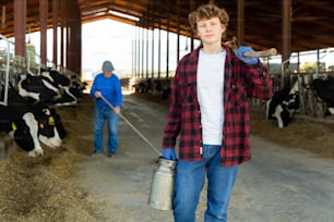 Confident teenage farm worker walking through cowshed after work on background of stall with cows, carrying pitchforks and milk can