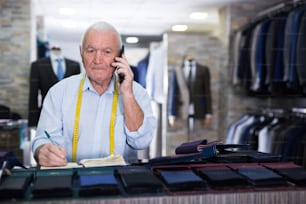 Professional tailor man talking on mobile phone and making notes in a notebook