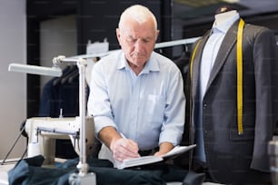Elderly man tailor uses notepad to record jacket measurements in workshop