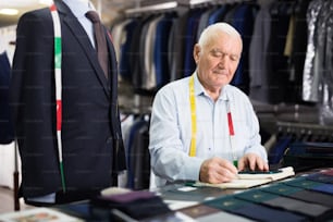 Elderly man tailor takes measurements from his jacket and writes them down in notebook in a workshop