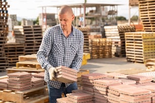 Jeune homme en gants en train de piquer des dalles de pavage sur des palettes dans un entrepôt de matériaux de construction