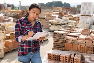 Concentrated hispanic woman worker controlling quantity of bricks at hardware store warehouse