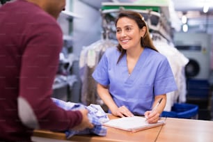 Cheerful woman laundry worker receiving dirty clothes from customer at dry-cleaning facility