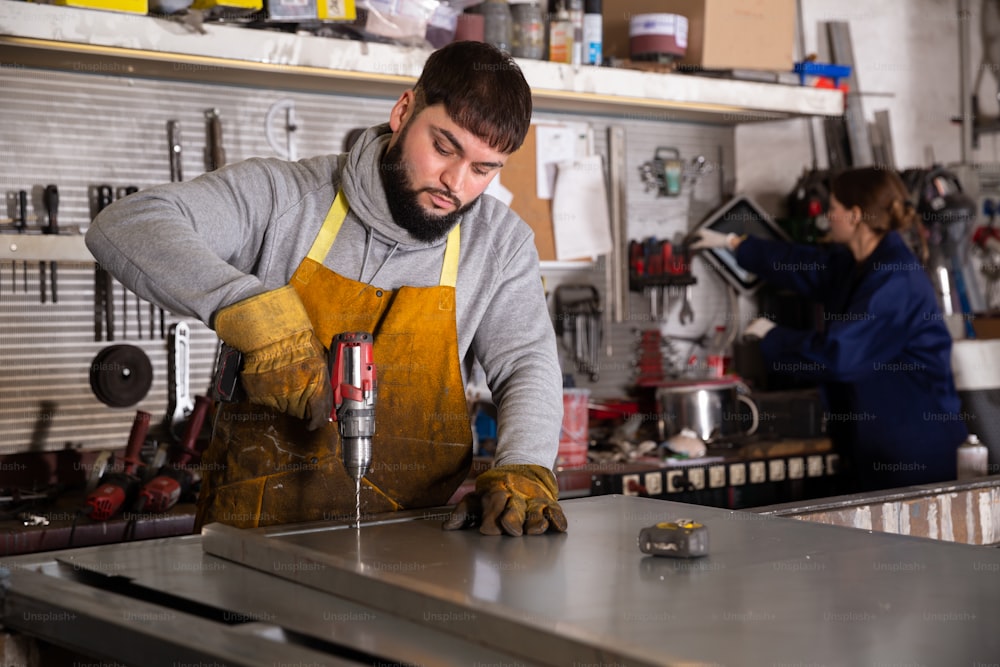 Joven artesano serio que trabaja con taladro eléctrico manual, perforando estructuras metálicas en un taller industrial