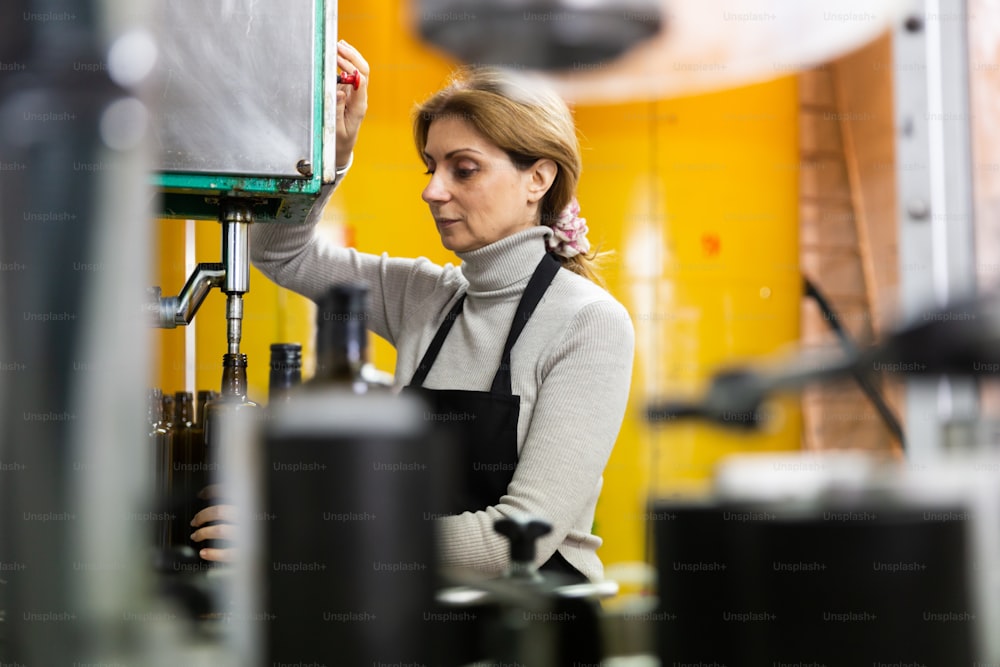 Femme travaillant sur un convoyeur d’embouteillage d’une usine de production alimentaire, remplissant des bouteilles en verre avec de l’huile d’olive