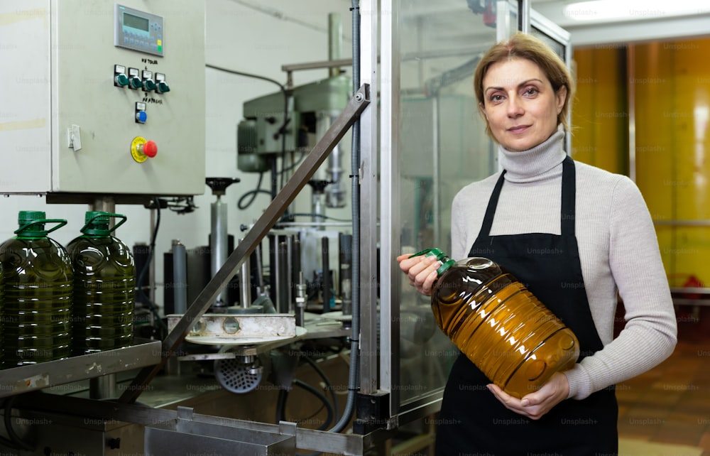 Femme prospère propriétaire d’une petite usine d’huile d’olive debout près d’une ligne d’embouteillage avec une bouteille en plastique remplie de produit fini dans les mains