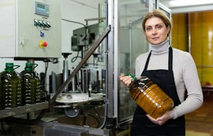 小さなオリーブオイル工場の成功した女性オーナーが瓶詰めラインの近くに立ち、完成品を手にしたペットボトルを持つ