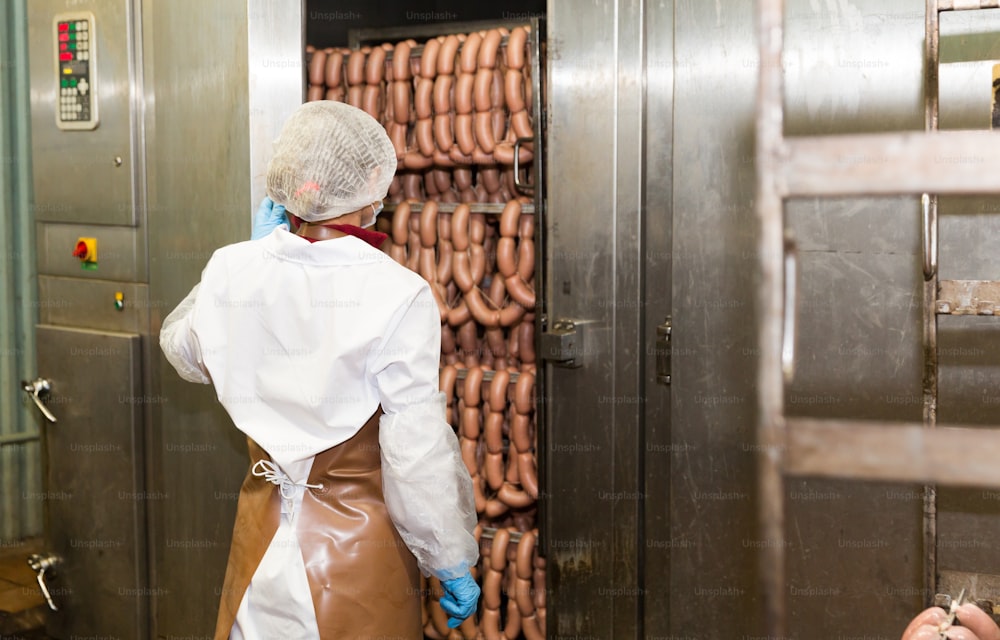 Female worker controlling hot processing of sausages in industrial smoking furnace at meat factory