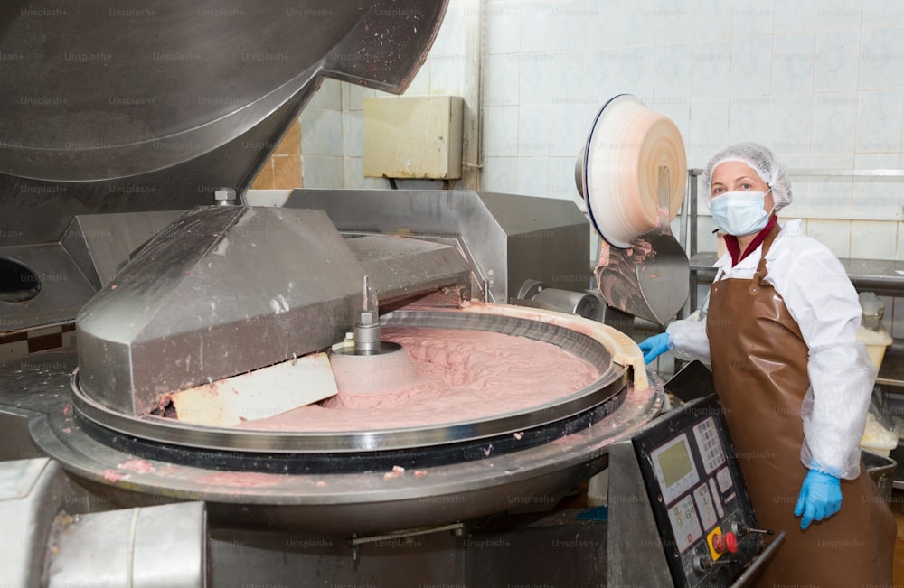 Arbeiterin, die den Prozess der Herstellung von fein gemahlenem Fleischteig in einer Schüssel, einem Zerkleinerer für Brühwurst und Würstchen in der Fleischfabrik kontrolliert