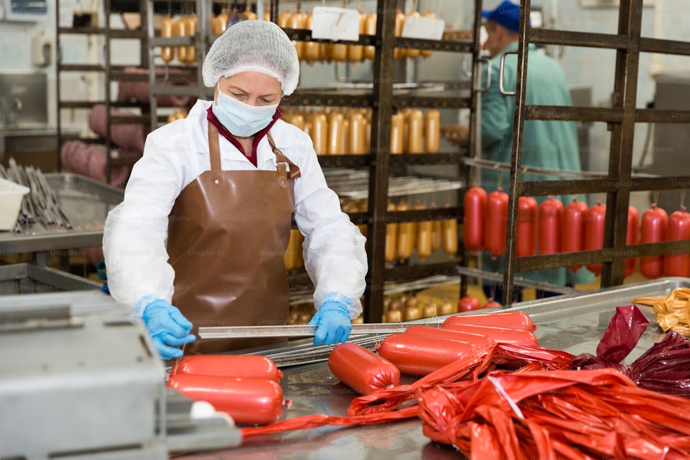 Focused woman hanging up raw sausages on rails for later hot processing at meat plant