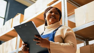 Female worker checking list of papers for inventory, working on stock logistics in warehouse. Young employee analyzing retail products to prepare merchandise distribution. Handheld shot.