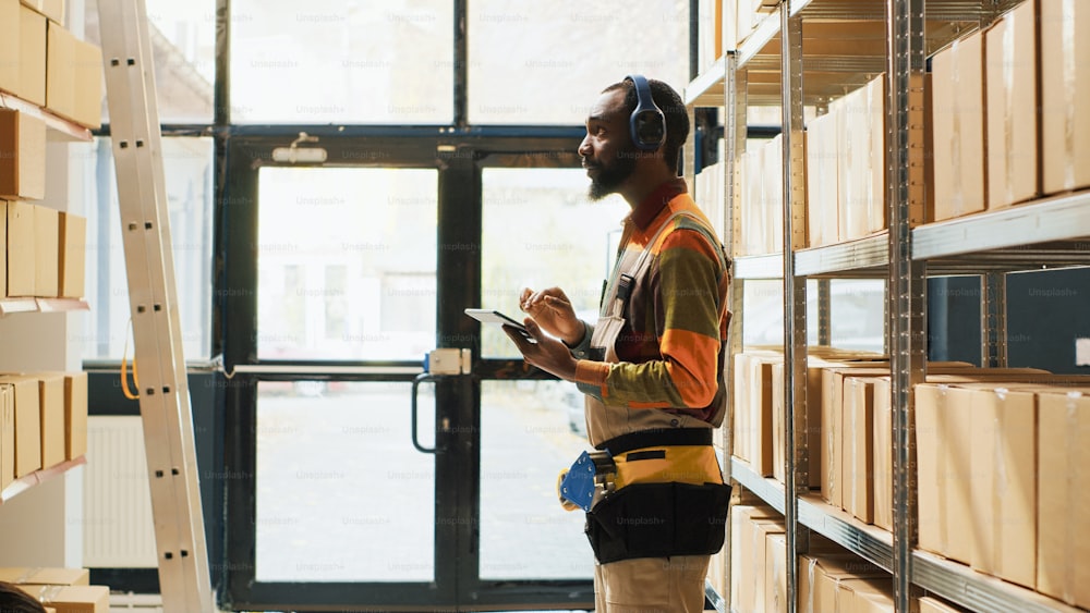 Happy young man listening to music checking boxes in warehouse space, enjoying songs on headset at work. Male worker singing and working on logistics and inventory with tablet. Tripod shot.