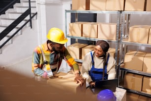 Distribution center workers checking merchandise inventory on laptop and packing freight for shipment. African american logistics managers working in storehouse full of cardboard boxes