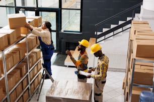 African american post office employee standing on ladder and taking parcel from shelf. All black package handlers team inspecting order and preparing cardboard box for shipment top view