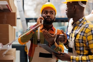 Warehouse logistics operator managing parcel dispatching on telephone. African american shipment managers asking about goods delivery in factory industrial storehouse on landline phone