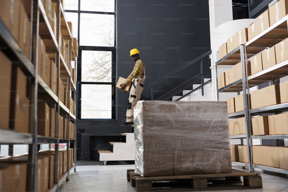 African american shipping company loader carrying heavy parcel downstairs. Post office man employee wearing protective helmet, holding cardboard box and going down to warehouse floor