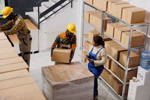 African american warehouse loader giving cardboard box to supervisor in post office storage room. Industrial storehouse woman manager inspecting freight packaging top view