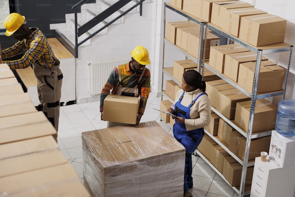 African american warehouse loader giving cardboard box to supervisor in post office storage room. Industrial storehouse woman manager inspecting freight packaging top view