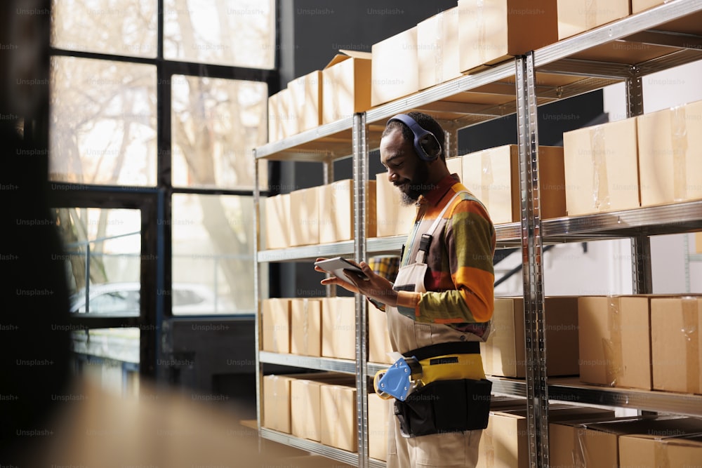 Warehouse manager in headphones doing inventory and keeping records in tablet software. African american industrial storehouse supervisor in earphones using stock management application