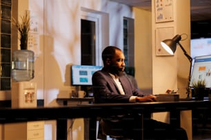 African american businessman standing at desk table analyzing financial graph on computer, working at company strategy late at night in startup office. Executive manager planning investment plan