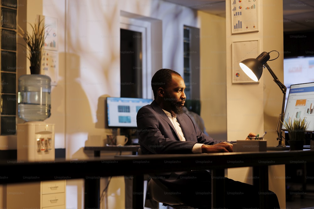 African american businessman standing at desk table analyzing financial graph on computer, working at company strategy late at night in startup office. Executive manager planning investment plan