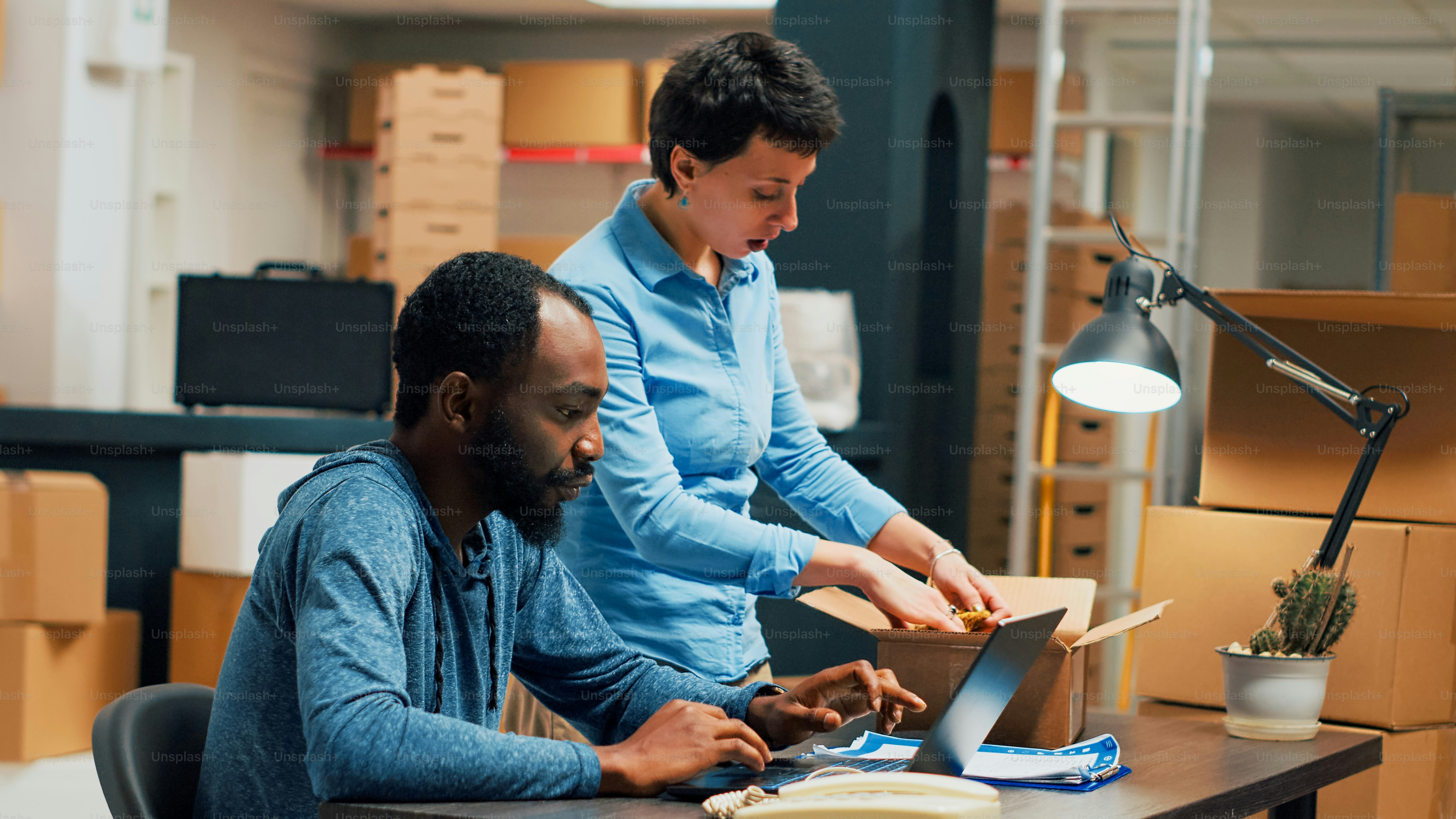Kleinunternehmer, die die Qualitätskontrolle des Vertriebs für die Startup-Entwicklung durchführen und mit dem Laptop im Lager an der Lieferung von Lagerbeständen arbeiten. Junge Leute, die Teamork machen, um Waren zu versenden. Aufnahme aus der Hand.