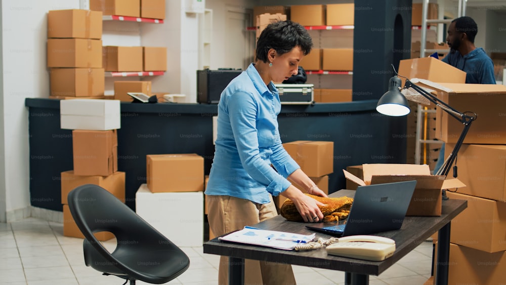 Small business owner planning order shipping with box, taking notes on laptop for stock inventory and working on delivering merchandise. Young adult selling products from warehouse racks.