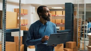 Male storage room worker using laptop to write products data, analyzing racks filled with cardboard packages for stock inventory. Young man planning logistics and merchandise management.